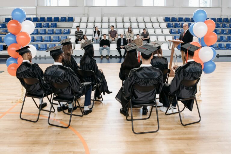 Graduates sitting in a gymnasium during a ceremony with colorful balloons.